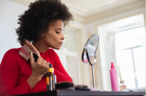Femme aux cheveux bouclés brossant ses cheveux devant un miroir, avec ses soins posés devant elle